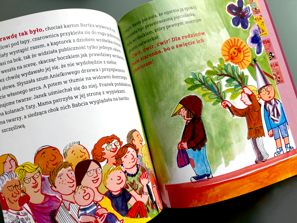 Przedszkolaki przygotowujące kostiumy do przedstawienia; Basia uśmiechnięta, trzyma w ręku rekwizyt, a nauczycielka pokazuje plan występu.