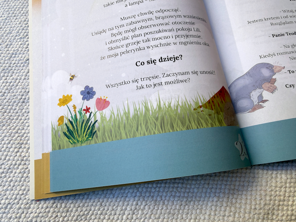 Książka „Przygody kłębuszka spod łóżka” otwarta na ilustrowanych stronach, przedstawiających przygody małego kłębuszka i dziewczynki Lili.