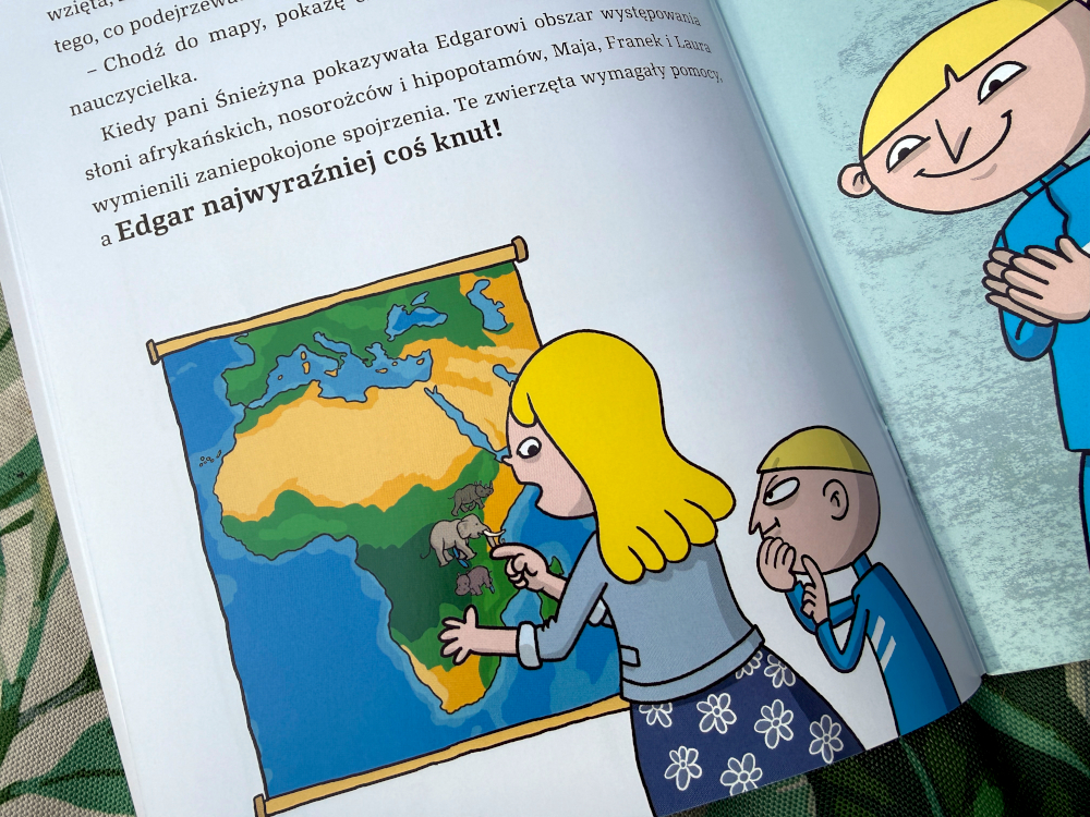Ilustracja z komiksu: nauczycielka wskazuje mapę Afryki z zaznaczonymi słoniami i nosorożcami, uczniowie obserwują
