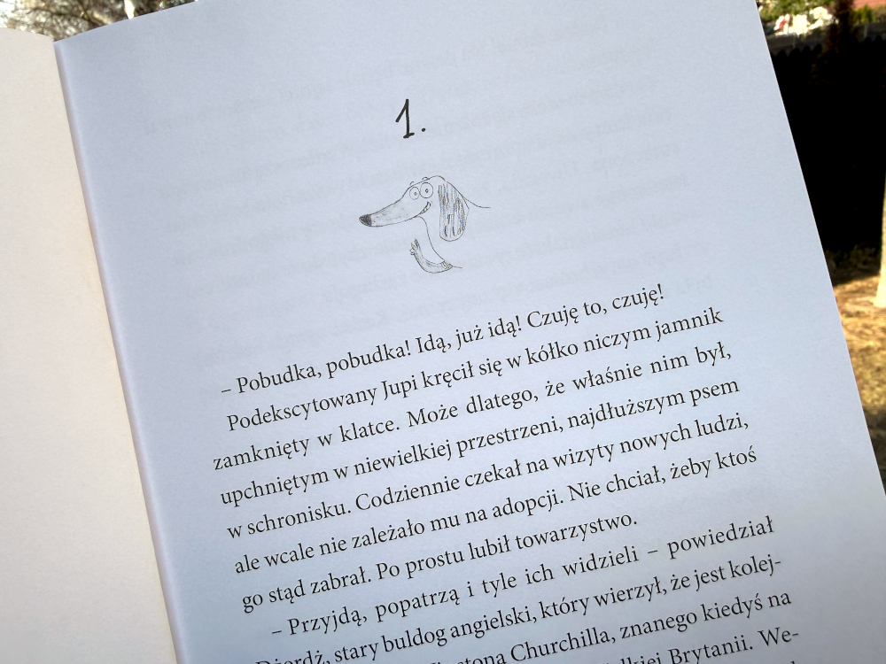 Otwarte strony książki z rozdziałem pierwszym i ilustracją jamnika Jupiego, widoczny tekst i minimalistyczna grafika