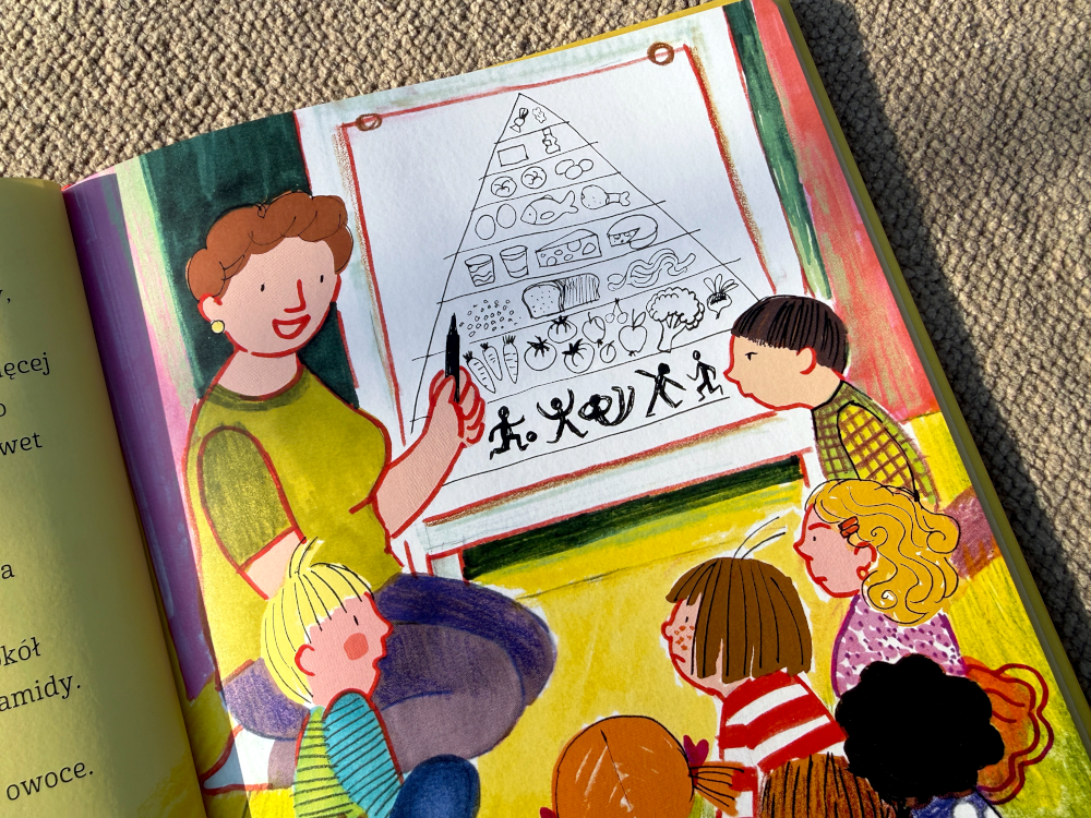 Scena z książki Basia i jedzenie – nauczycielka pokazuje dzieciom piramidę żywieniową na tablicy podczas zajęć w przedszkolu