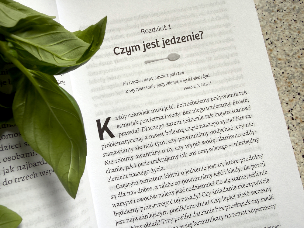 Otwarte strony książki Jak rozmawiać z dziećmi o jedzeniu z rozdziałem o roli jedzenia, obok zielone liście bazylii