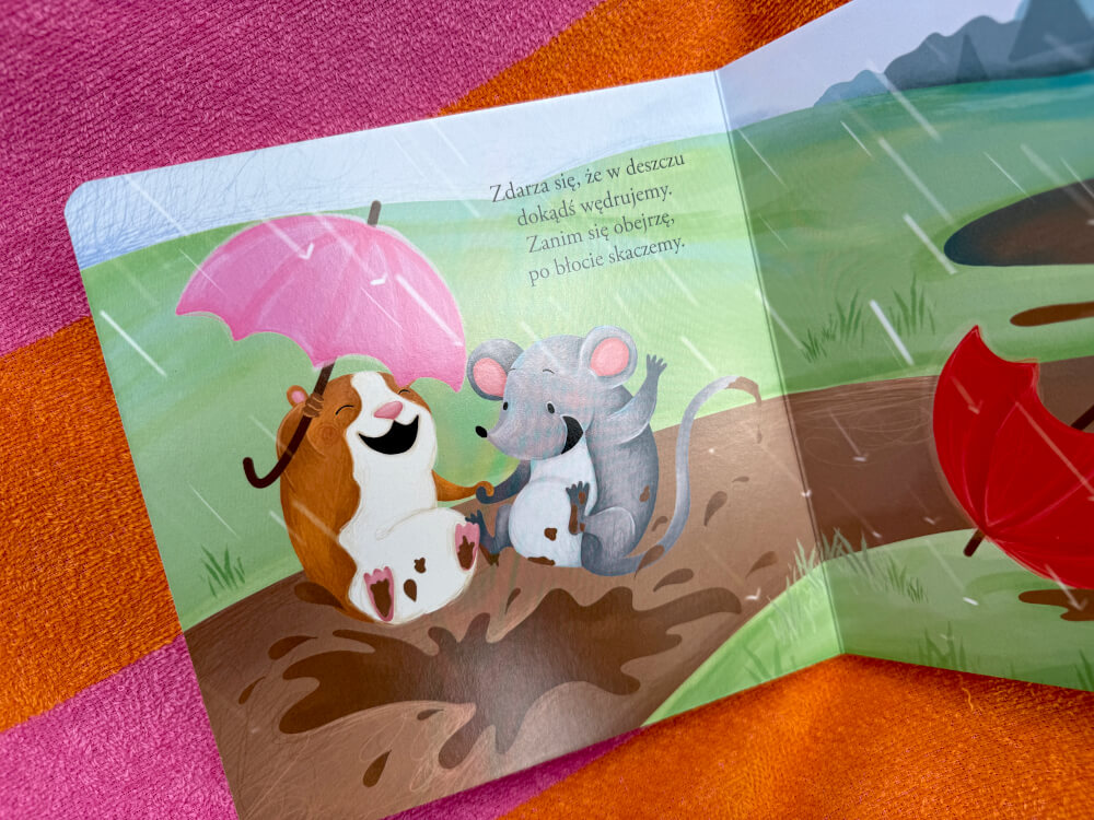 Ilustracja przedstawiająca dwie postacie z różowym parasolem radośnie bawiące się i siedzące w błotnistych kałużach.