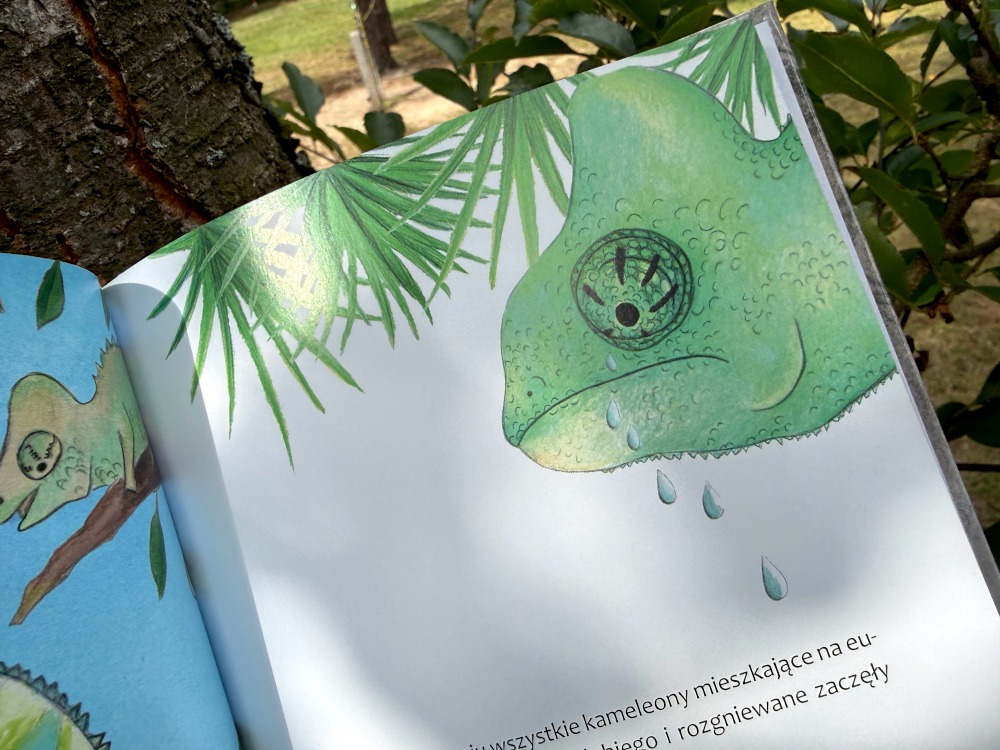 Zbliżenie na zmartwionego, płaczącego kameleona z ilustracji. Widoczne duże łzy spływające po zielonej skórze. W dole fragment tekstu o kameleonach.