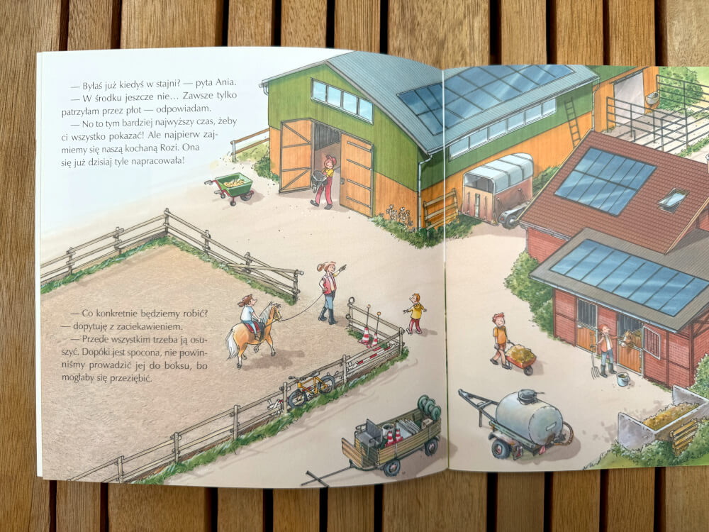 Ilustracja przedstawiająca podwórko stajni, budynki z solarnymi dachami oraz postacie przygotowujące konie do jazdy z książki