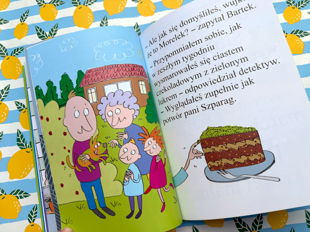 Ilustracja na stronie książki przedstawia bohaterów w ogrodzie. Detektyw Zagadka trzyma psa, a obok na talerzu stoi kawałek ciasta z zielonym kremem.