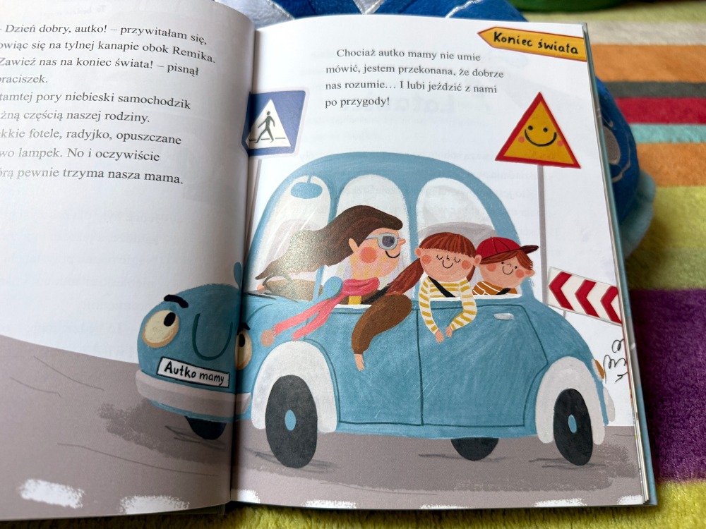 Ilustracja w książce Autko mamy przedstawiająca rodzinę podróżującą radosnym, niebieskim samochodem z dziećmi.